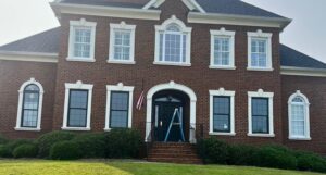 Front exterior before view of a large two-story red brick house, showing the original white window trim and white architectural moldings prior to being updated.