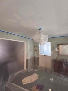 Interior ceiling painting preparation in Columbia, SC, showing a dining room with walls and furniture meticulously covered in protective plastic sheeting and a chandelier wrapped for safety.